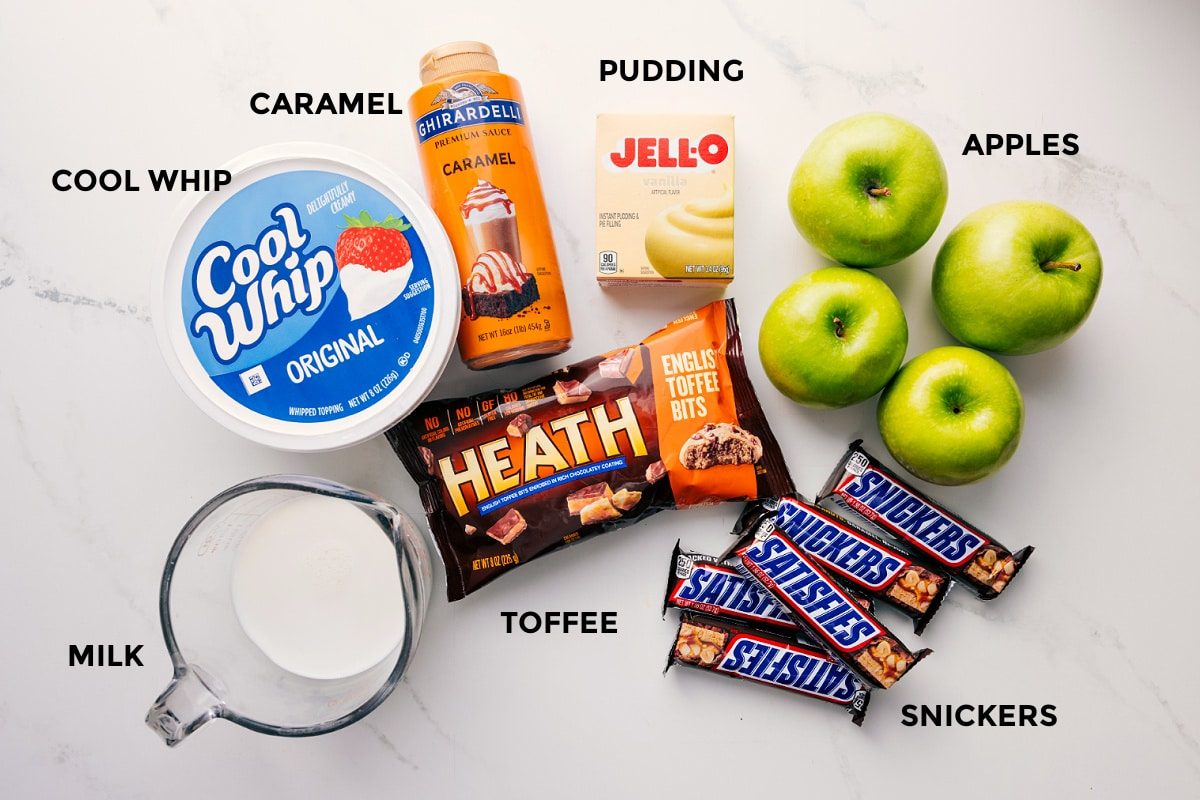 All the ingredients in this recipe prepped out for straightforward assembly including the milk, cool whip, caramel, toffee, snickers, apples, and jello.