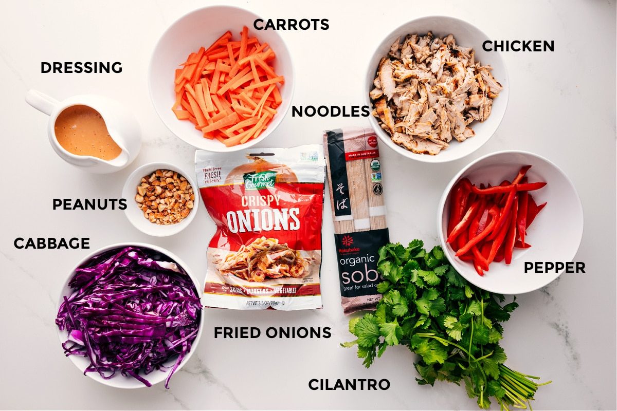 All the ingredients in this recipe prepped out for straightforward assembly including the dressing, peanuts, cabbage, fried onions, carrots, noodles, chicken, pepper, and cilantro.