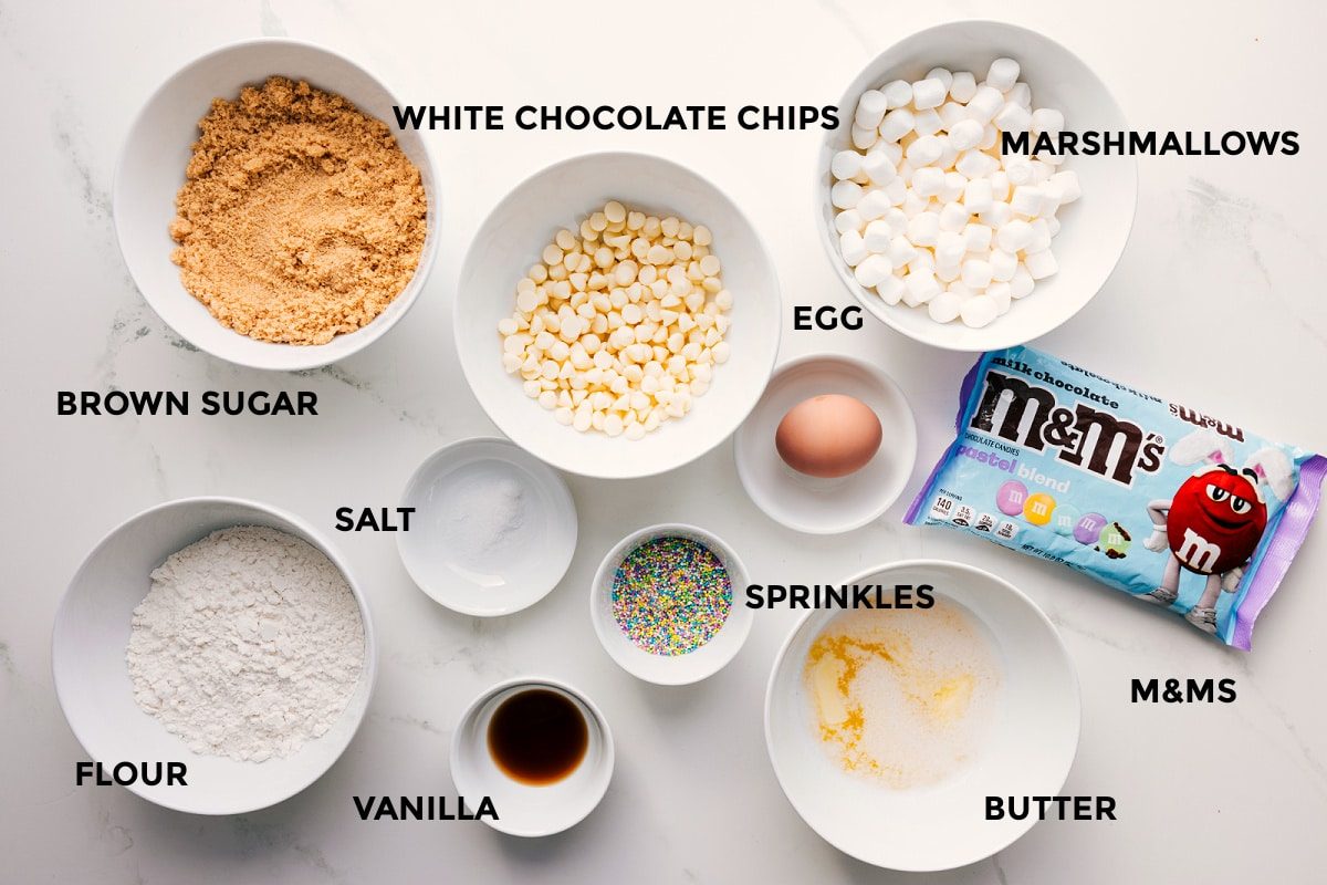 All the ingredients in this recipe prepped out for straightforward assembly including the brown sugar, flour, vanilla, salt, white chocolate chips, marshmallows, egg, M&Ms, butter, and sprinkles.