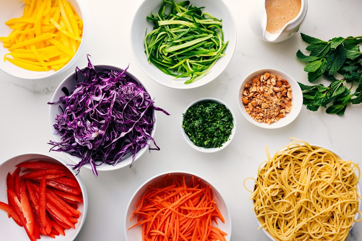 All the ingredients in this recipe prepped out for straightforward assembly including the peppers, cabbage, carrots, pasta, peanuts, dressing, herbs, and cucumbers.