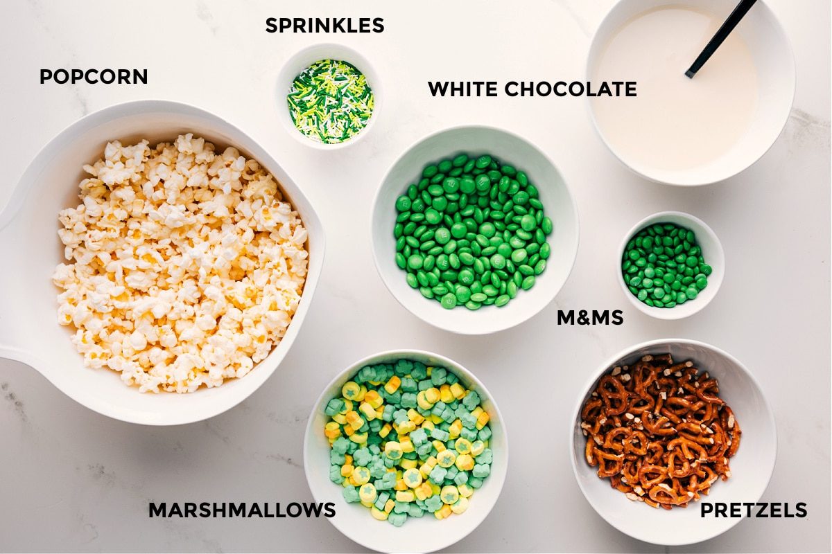 All the ingredients for this recipe prepped out for straightforward assembly including the popcorn, sprinkles, marshmallows, M&Ms, white chocolate, and pretzels.