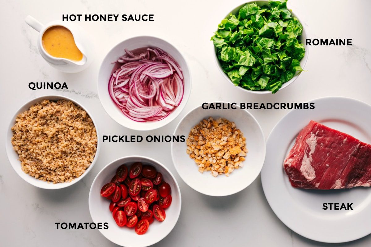 All the ingredients in this recipe prepped out for straightforward assembly including the hot honey sauce, quinoa, tomatoes, pickled onions, romaine, garlic breadcrumbs, and steak.