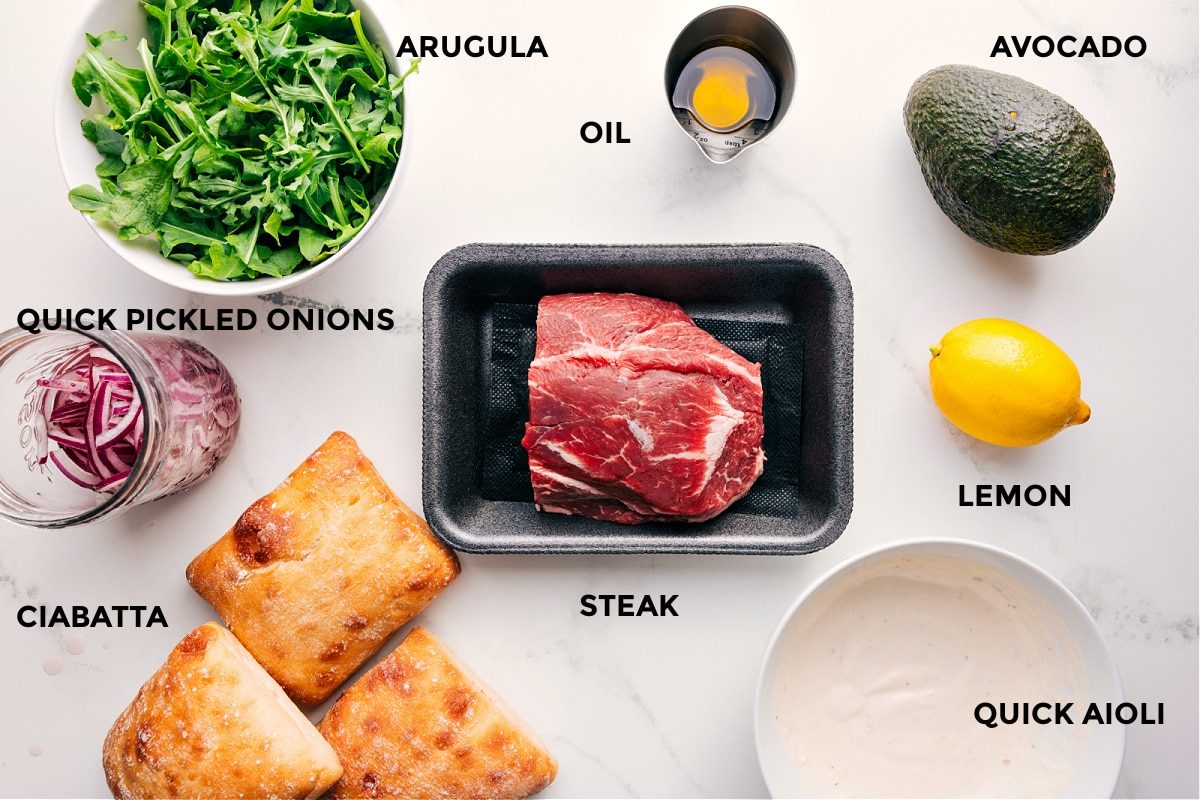 All the ingredients in this recipe prepped out for straightforward assembly including the arugula, pickled red onions, ciabatta, steak, oil, avocado, lemon, and a quick aioli.