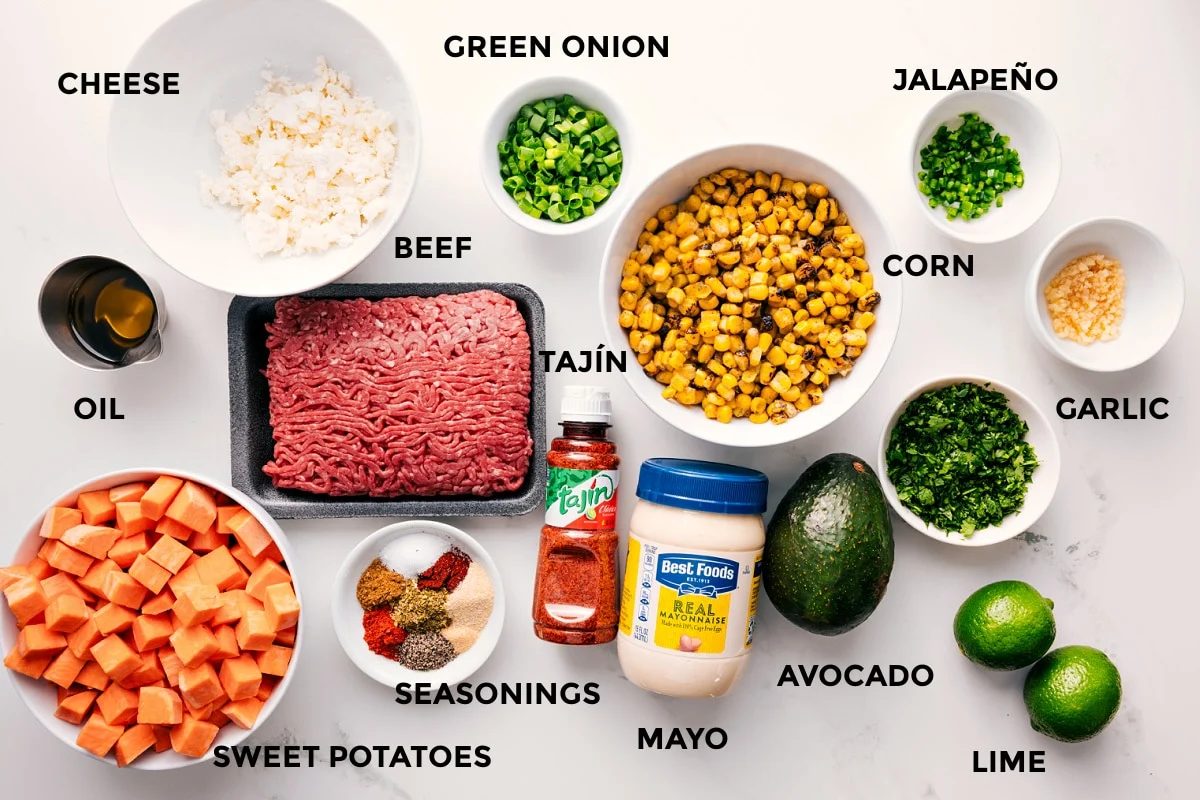 All the ingredients in this recipe prepped out for straightforward assembly including the cheese, oil, sweet potatoes, beef, seasonings, tajin, mayo, avocado, lime, garlic, jalapeno, corn, and green onions.