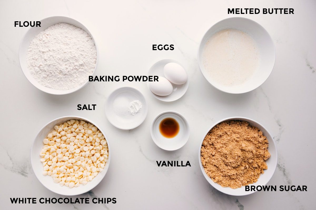 All. the ingredients in this recipe prepped out for straightforward assembly including the flour, white chocolate chips, salt, baking powder, eggs, vanilla, melted butter, and brown sugar.