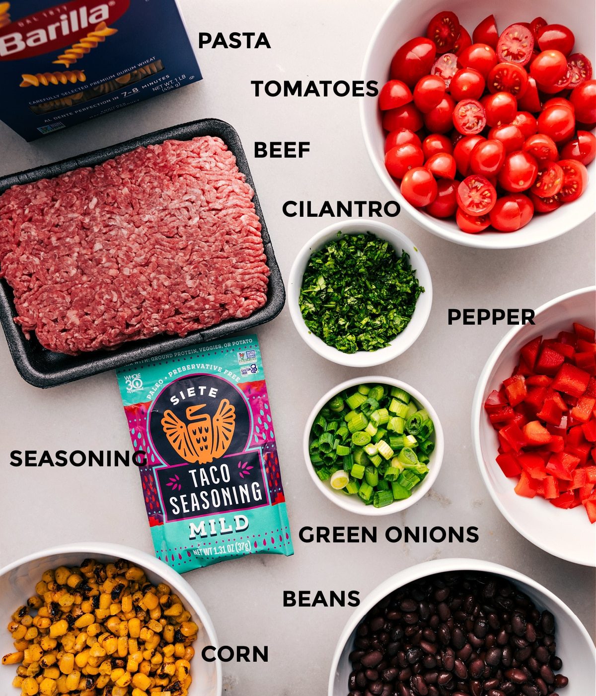 All the ingredients—pasta, meat, tomatoes, cilantro, pepper, green onions, seasoning, beans, and corn—are prepped and ready for straightforward assembly.