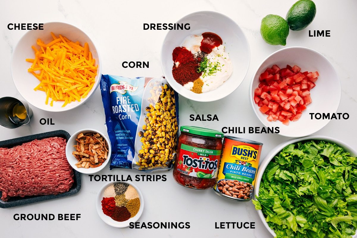 All the ingredients in this recipe prepped out for straightforward assembly including the ground beef, oil, cheese, tortilla strips, seasonings, salsa, chili beans, lettuce, tomato, lime, and dressing.