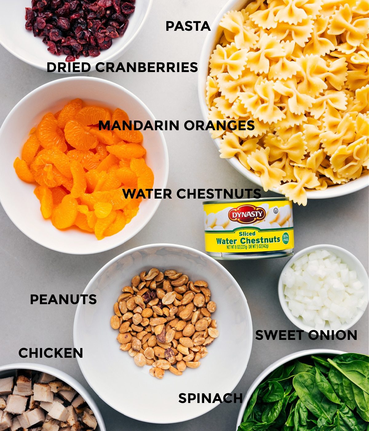 Ingredients including dried cranberries, pasta, water chestnuts, oranges, onion, spinach, peanuts, and chicken prepped out for straightforward assembly.