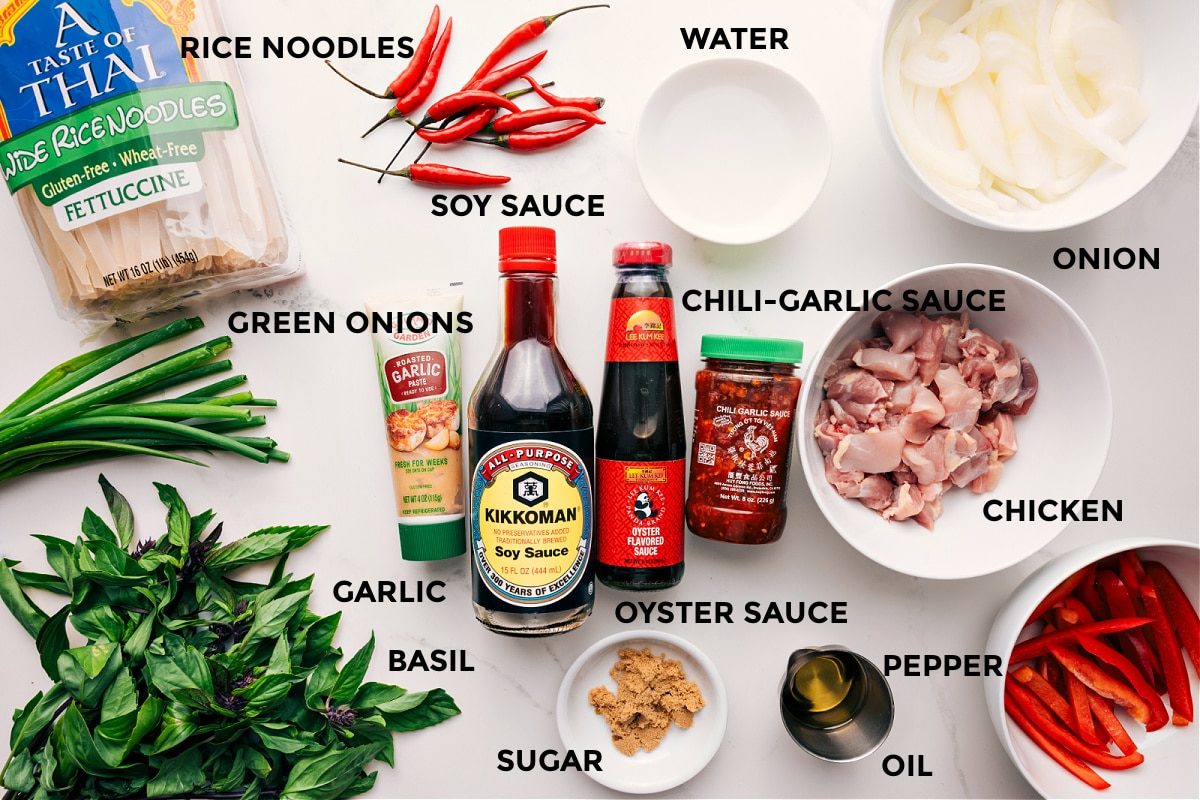 All the ingredients in this recipe prepped out for straightforward assembly including the rice noodles, green onions, garlic, basil, soy sauce, chili garlic sauce, oyster sauce, sugar, oil, pepper, chicken, onion, and water.