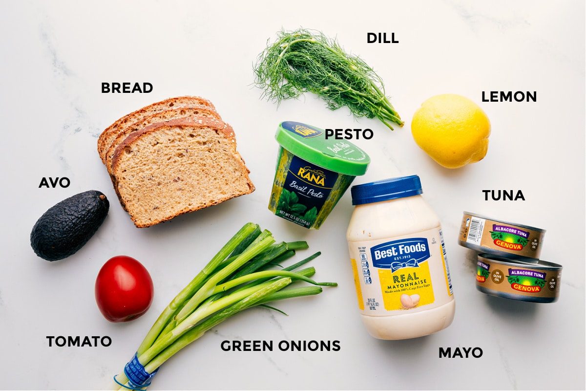 All the ingredients prepped for straightforward assembly, including avocado, tomato, bread, green onions, pesto, dill, lemon, tuna, and mayo.