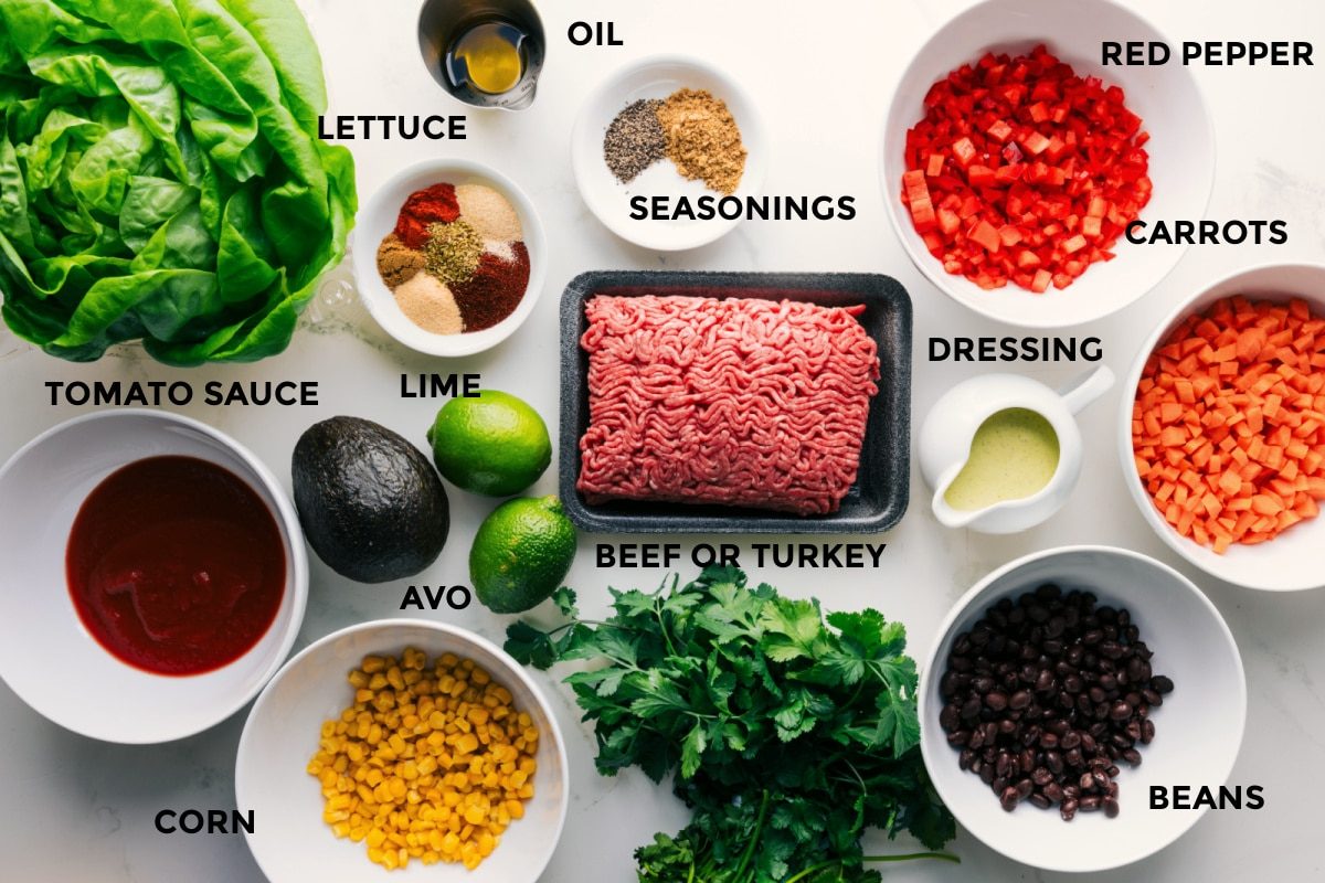 All the ingredients in this recipe prepped out for straightforward assembly including the lettuce, tomato sauce, corn, avocado, seasonings, limes, beef or turkey, beans, dressing, carrots, red pepper, and oil.