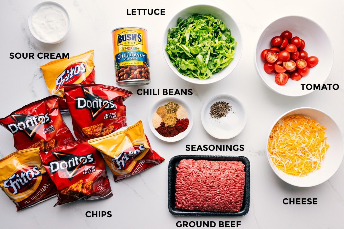 Ingredients in this recipe prepped for straightforward assembly, including sour cream, chili beans, lettuce, tomato, cheese, seasonings, ground beef, and chips.