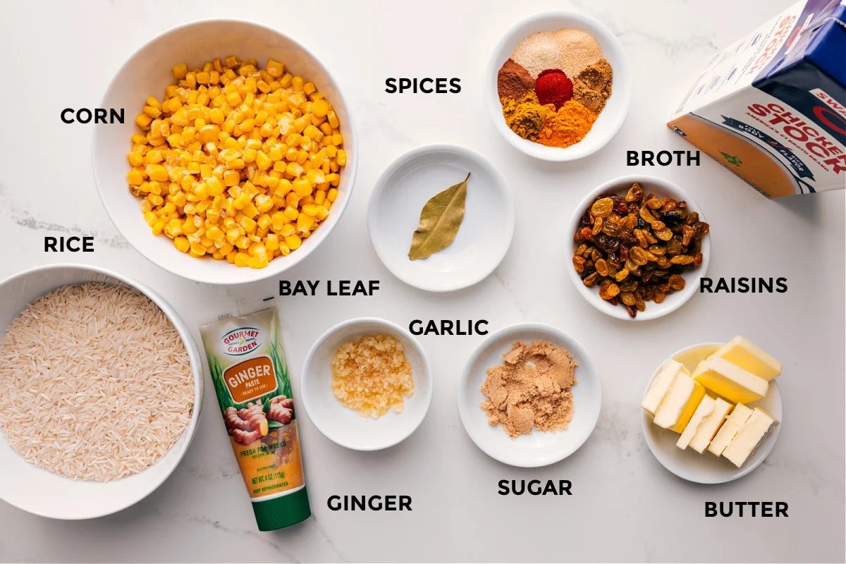 All the ingredients in this recipe prepped out for straightforward assembly including the rice, corn, ginger, bay leaf, garlic, sugar, butter, raisins, broth, and spices.