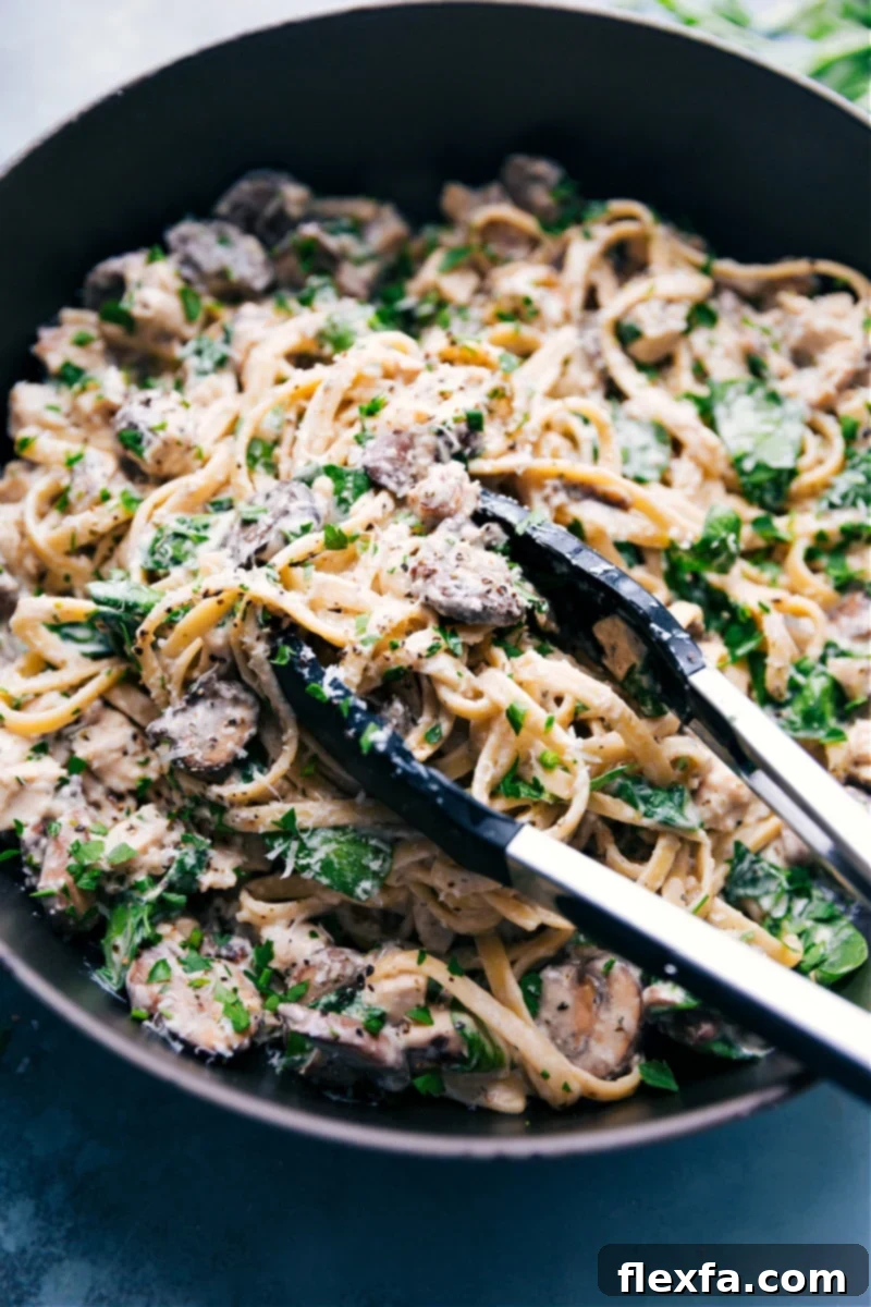 A large skillet filled with creamy chicken pasta, featuring fettuccine, chicken pieces, mushrooms, and spinach, with a serving spoon lifting a generous portion.