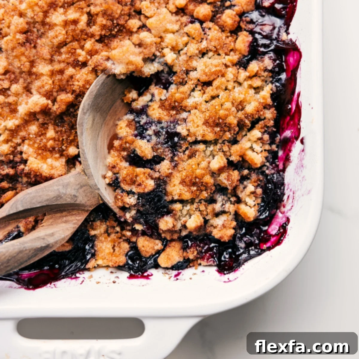 A freshly baked, golden-brown blueberry cobbler, still warm in its ceramic dish, with a serving spoon poised to scoop out a portion.