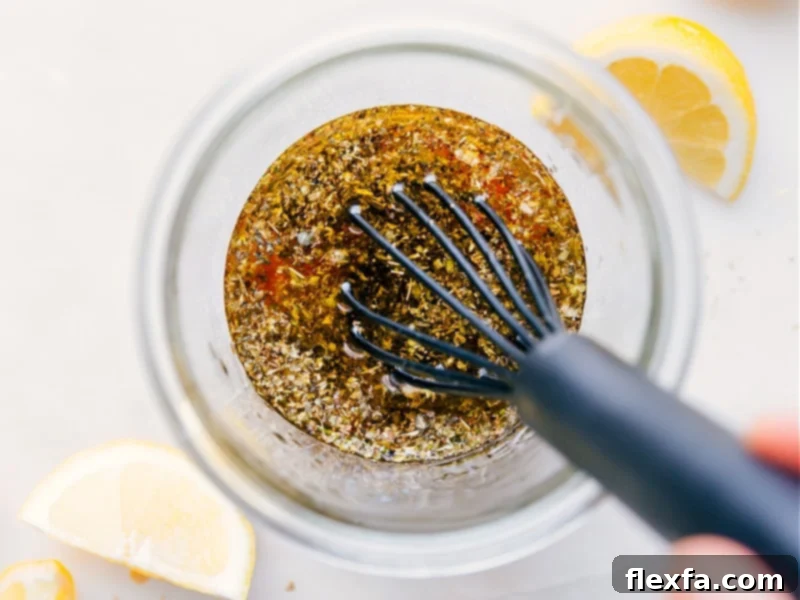 A whisk vigorously blending the ingredients for Greek Salad Dressing in a clear glass bowl, showcasing the emulsification process as the liquids come together beautifully.