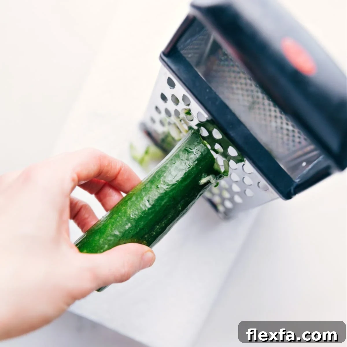 A hand grating an English cucumber directly into a bowl lined with paper towels to prepare for squeezing out excess liquid for Tzatziki Sauce.