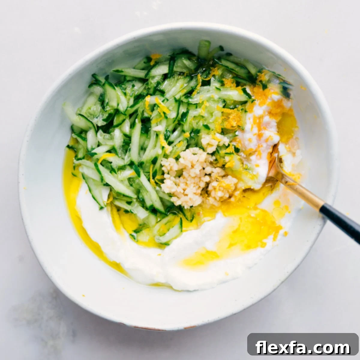 All the Tzatziki Sauce ingredients, including grated cucumber, yogurt, lemon, garlic, herbs, olive oil, and red wine vinegar, are combined in a bowl and being whisked together.