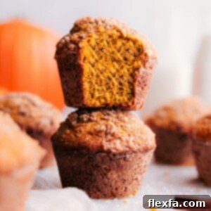 A stack of pumpkin muffins with a warm, golden tone, one at the front displaying a bite taken, revealing the moist interior.