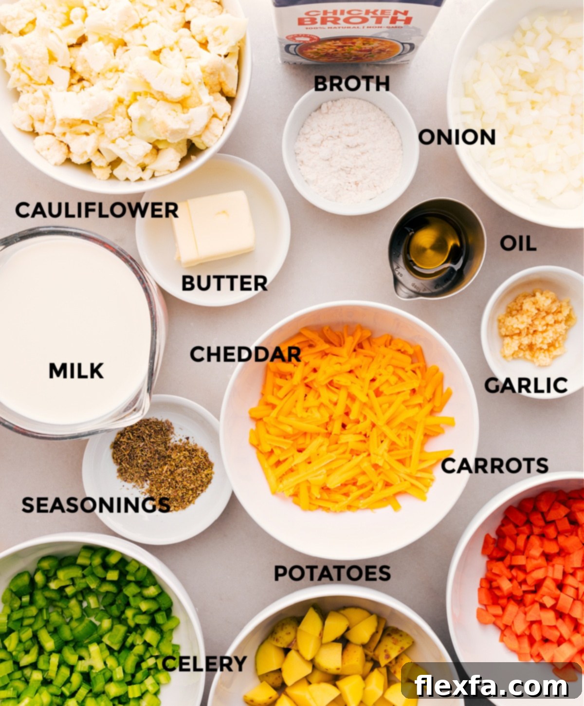 A beautiful arrangement of fresh ingredients for making creamy cauliflower soup: blocks of cheddar, colorful carrots, celery, onions, garlic, broth, butter, milk, and a whole head of cauliflower.