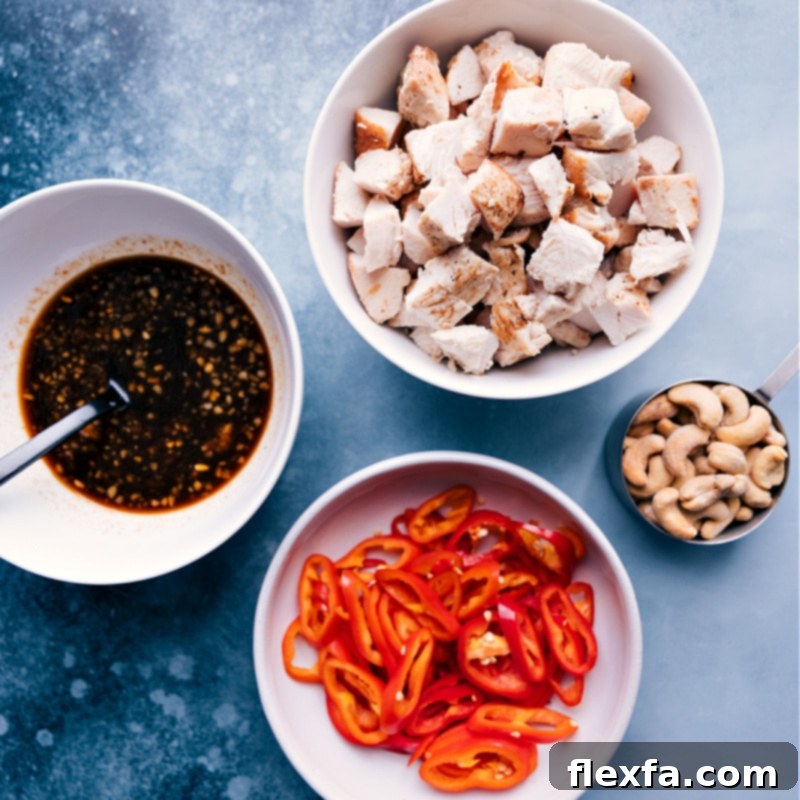 Carefully arranged ingredients for Cashew Chicken: cooked chicken, dry-roasted cashews, thinly sliced red bell peppers, and a bowl of the prepared savory sauce.
