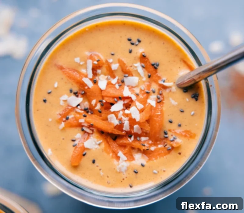 A beautifully presented Carrot Smoothie in a clear glass, garnished generously with fresh shredded coconut, chia seeds, and a sprinkle of grated carrots, showcasing its rich texture and appealing toppings.