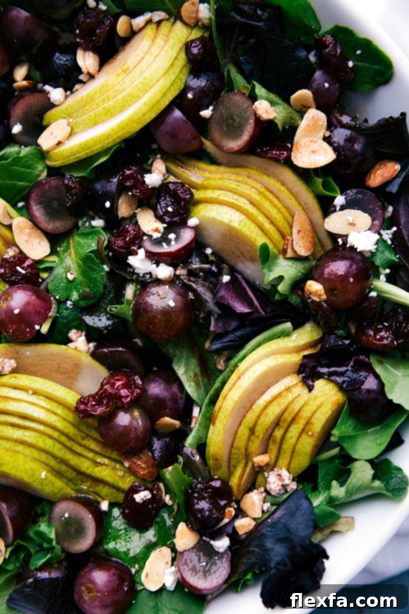 A beautifully arranged side dish, showcasing a vibrant medley of colorful components: mixed greens, red grapes, pear, almonds, dried cherries, and crumbled feta, all perfectly dressed and ready to enjoy.