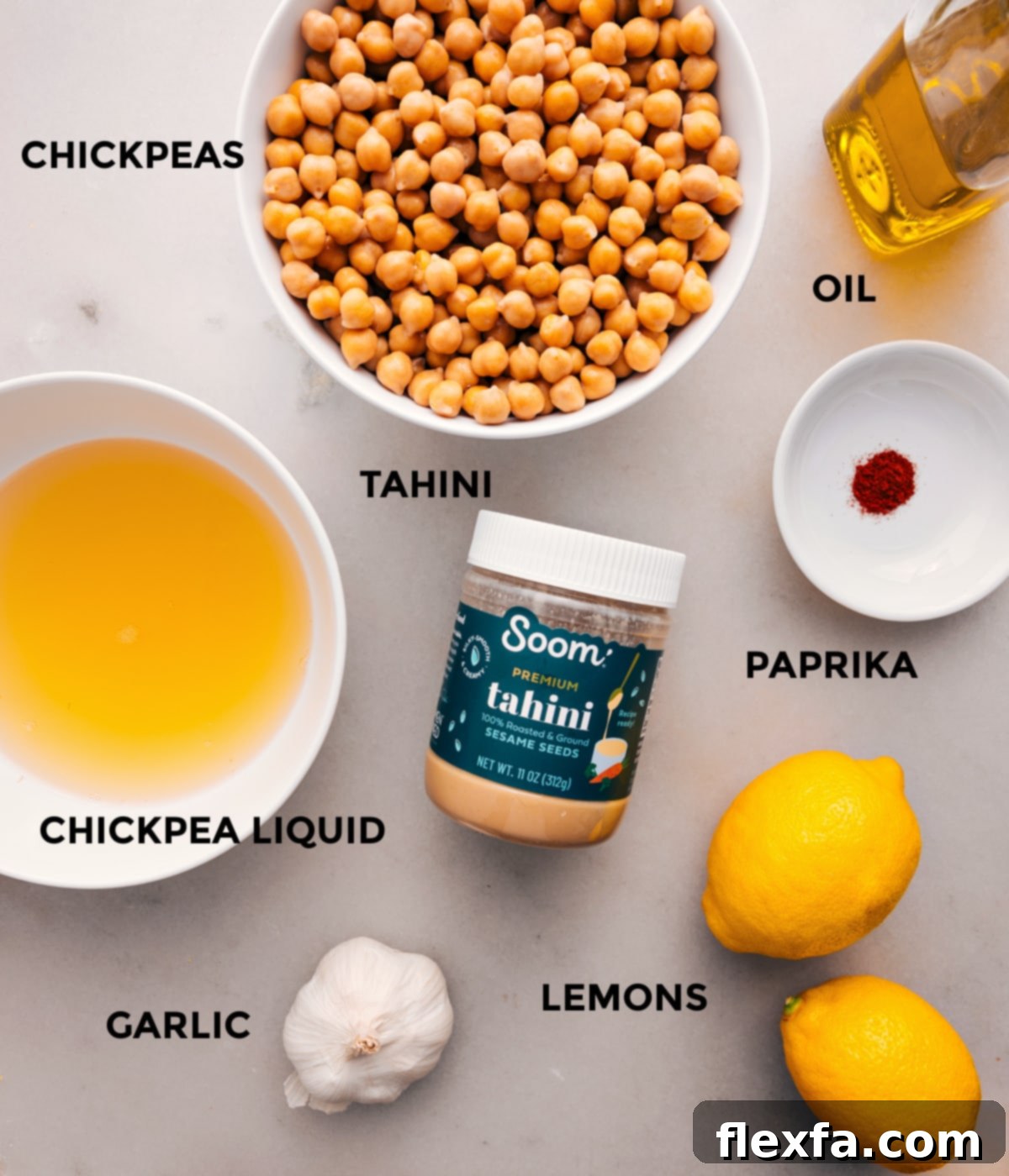 All the essential ingredients for making hummus are neatly prepped and arranged, including a bottle of olive oil, two cans of chickpeas, a jar of tahini, vibrant paprika, fresh lemons, and whole garlic cloves.