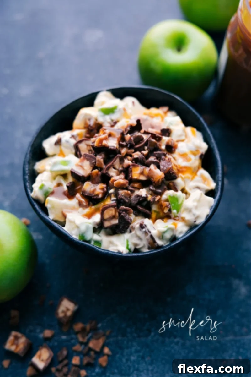 A vibrant Snickers Salad in a clear bowl, featuring chunks of Snickers candy bars, diced green apples, and a creamy white base. A spoon is partially submerged, ready for serving.