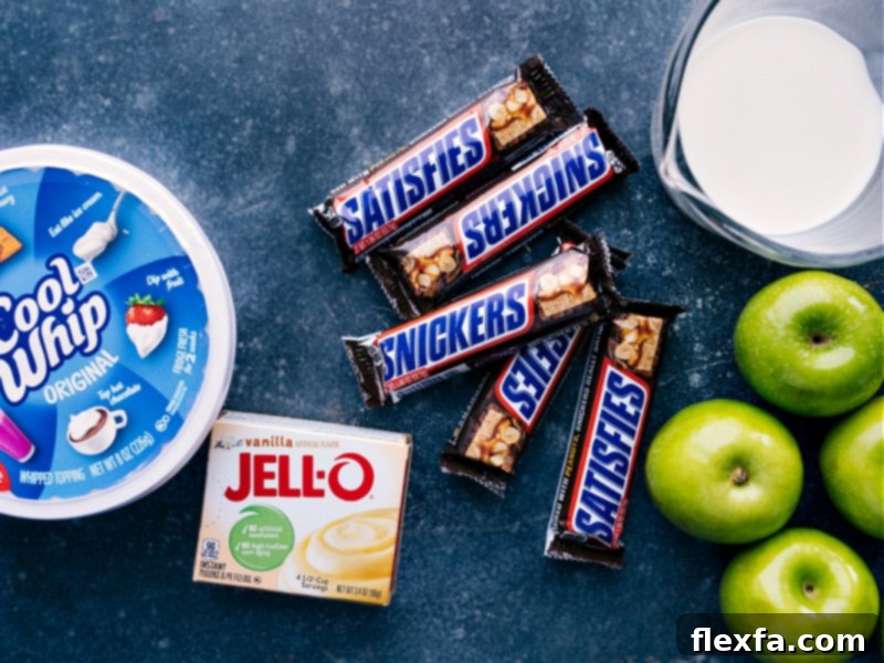 An overhead shot showcasing all the fresh ingredients for Snickers Salad: diced Snickers bars, chopped Granny Smith apples, a package of instant vanilla pudding mix, a carton of milk, and a tub of frozen whipped topping.
