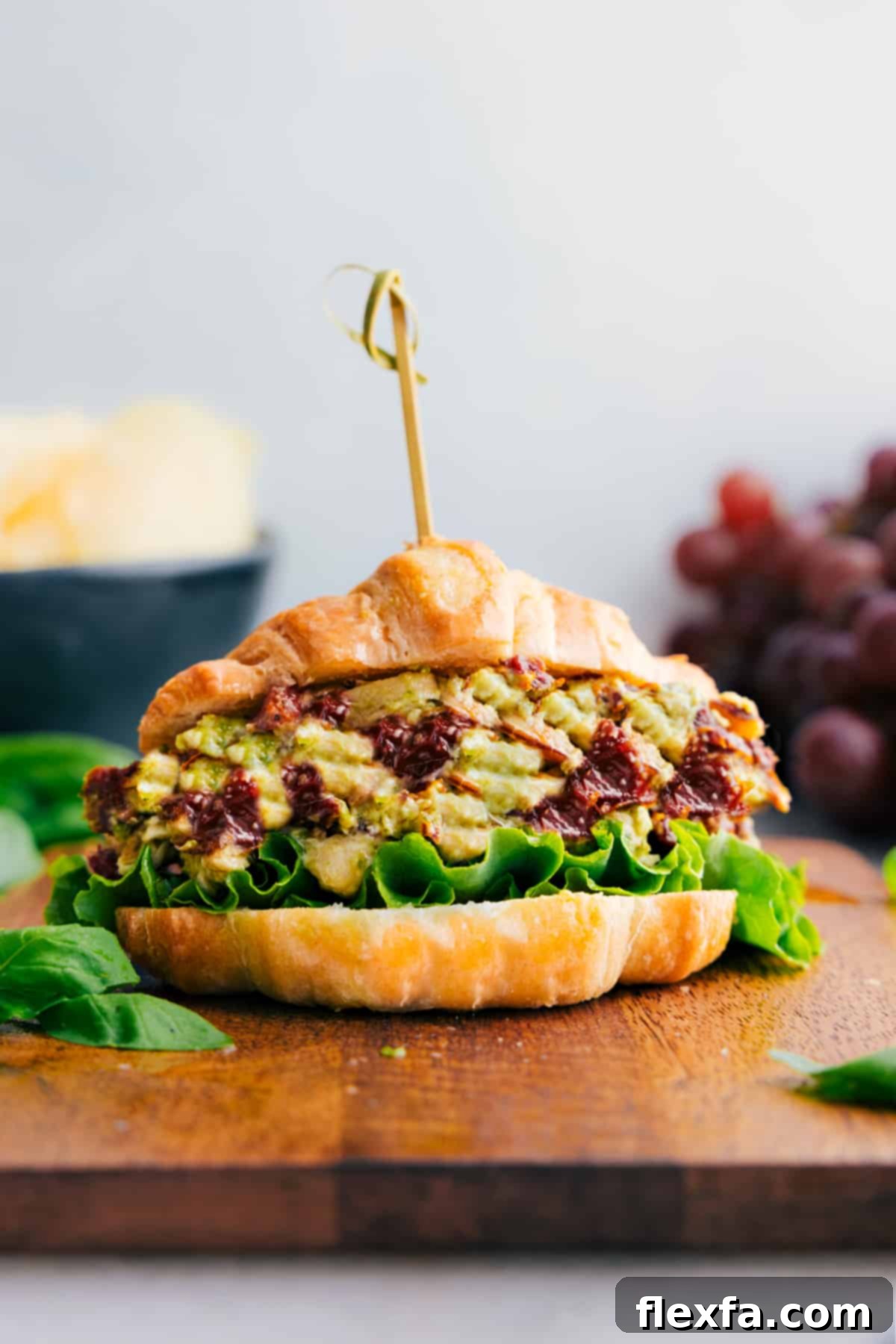 Sandwich with pesto chicken salad in side; grapes and chips in background, showcasing a fresh and vibrant meal.