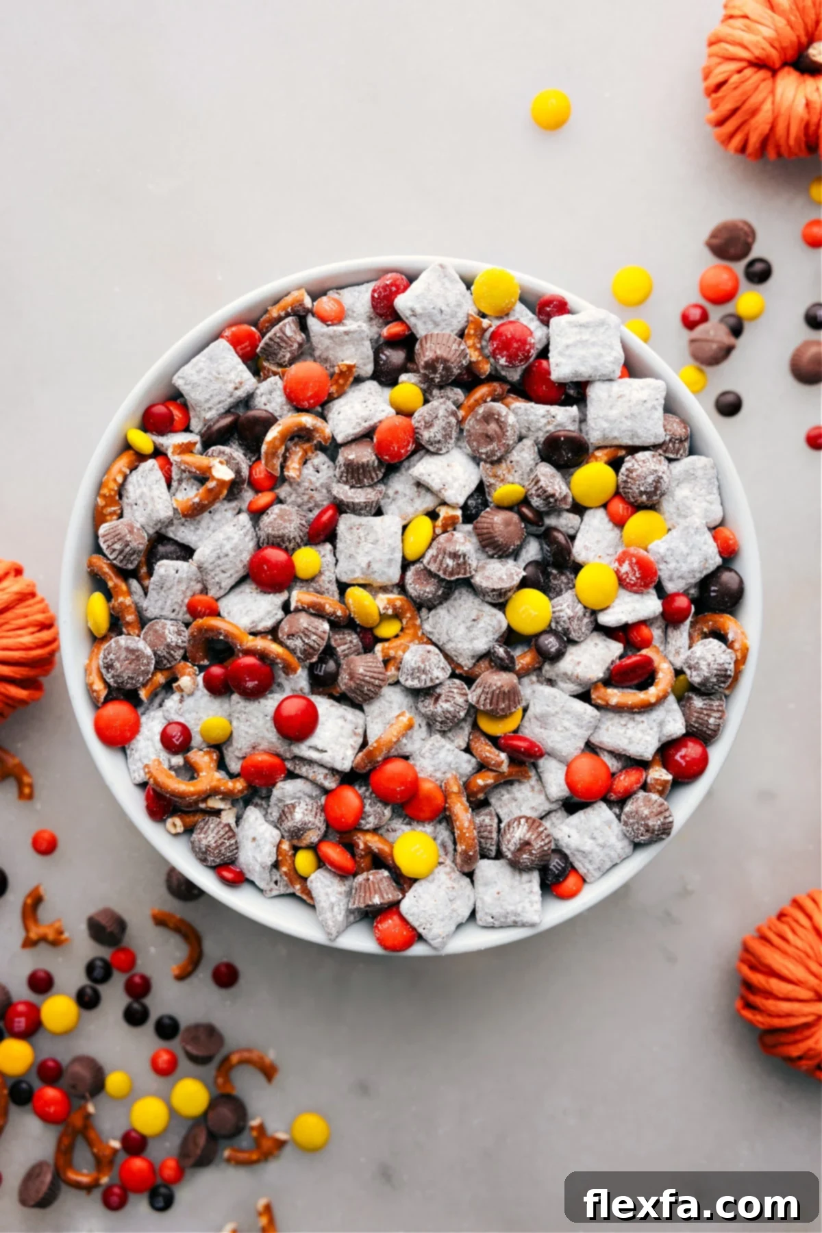 A vibrant bowl of Fall Muddy Buddies, featuring chocolate and powdered sugar-coated Chex cereal, adorned with colorful autumn candies like M&M's and mini Reese's, along with crunchy pretzel pieces, ready to be enjoyed as a festive fall snack.