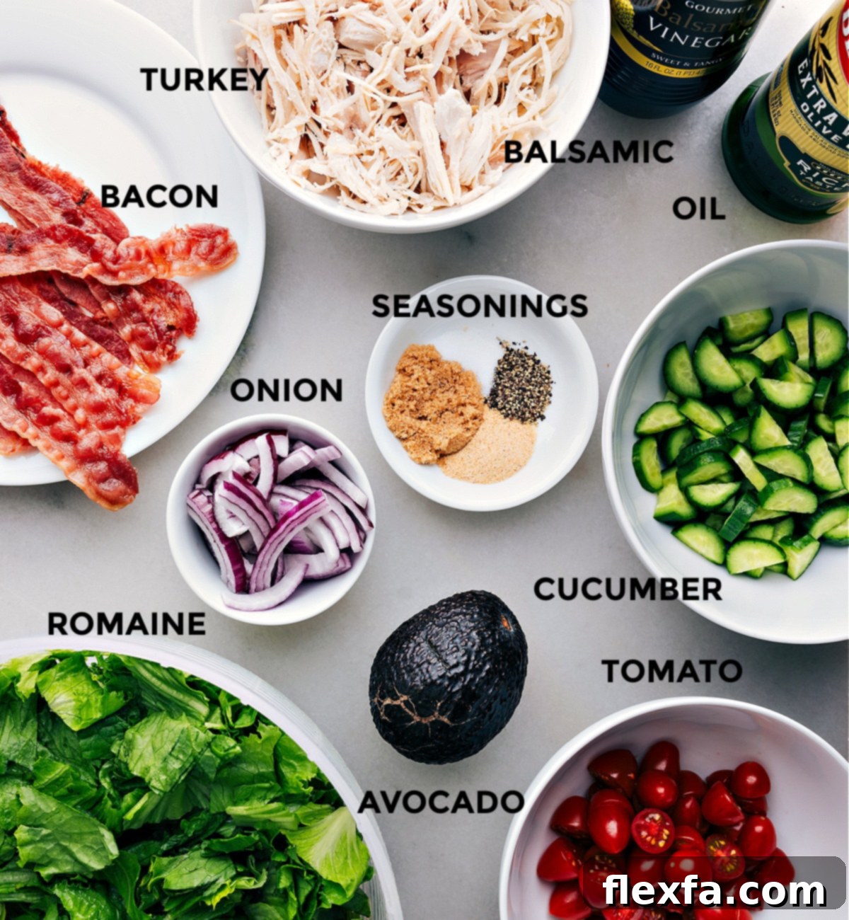 Ingredients In Turkey Bacon Avocado Salad All fresh ingredients for Turkey Bacon Avocado Salad spread out on a counter, including lettuce, tomatoes, cucumbers, onion, avocado, cooked turkey, and bacon, ready for assembly.