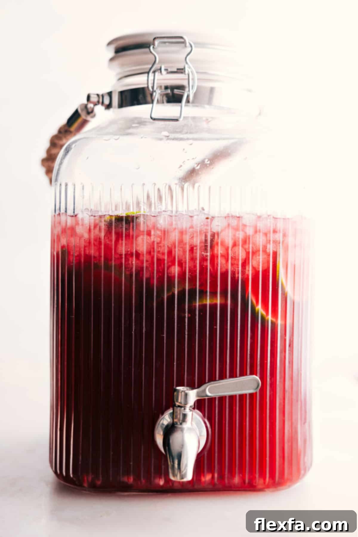 A large clear drink dispenser filled with festive Christmas punch, adorned with lime and pomegranate garnishes, ready for self-service at a party.