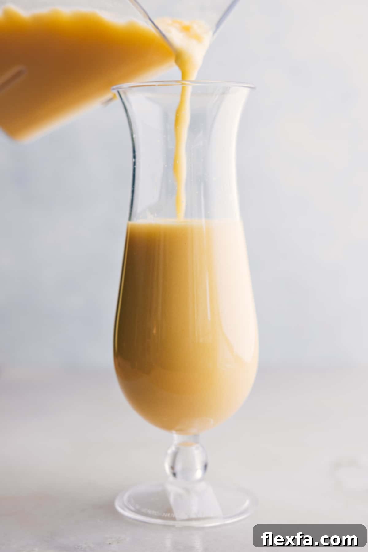 The Best Pina Colada Being Poured A freshly blended Pina Colada being poured from a blender into a tall hurricane glass, highlighting its creamy, frosty texture.