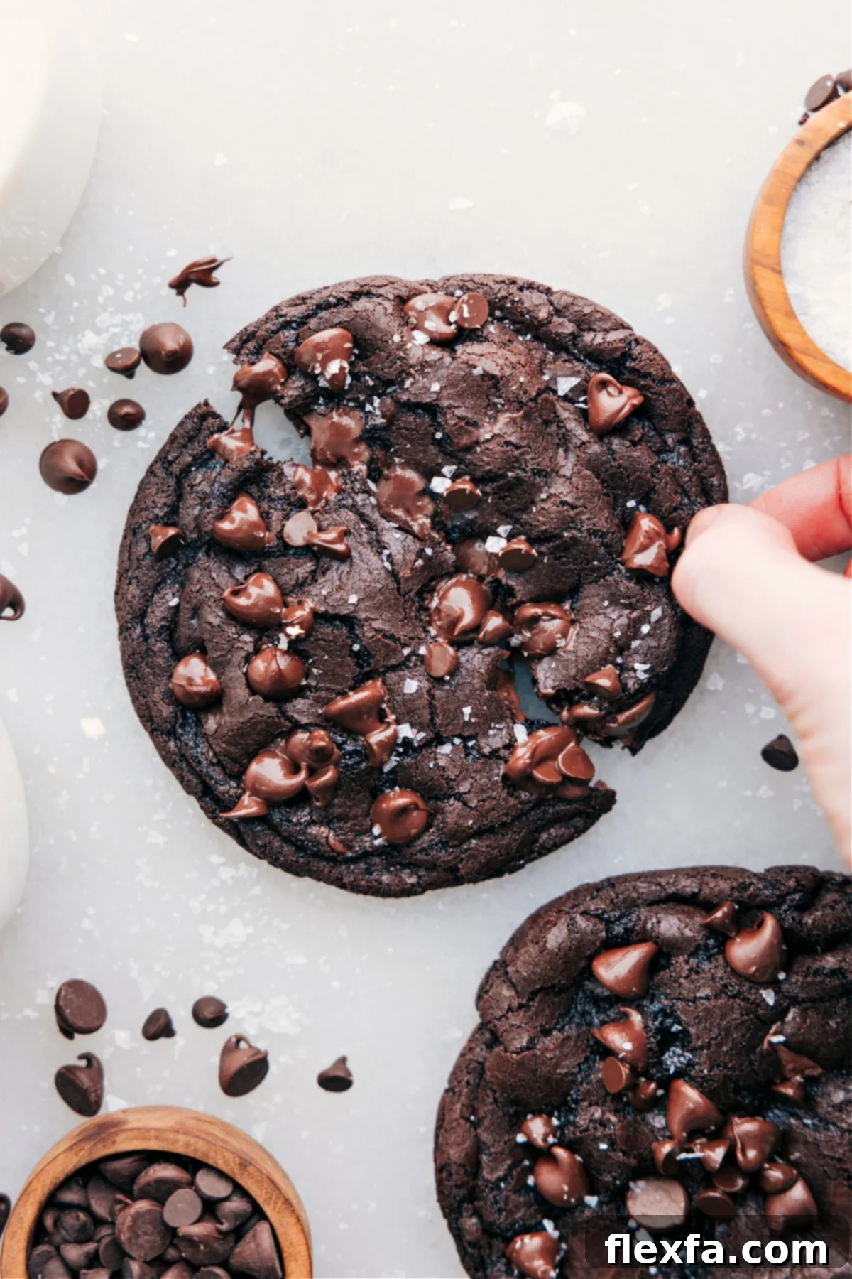 A single serve double chocolate chip cookie topped with flaky sea salt, being broken open by a hand to reveal its wonderfully gooey, melty chocolate center, emphasizing its irresistible texture.