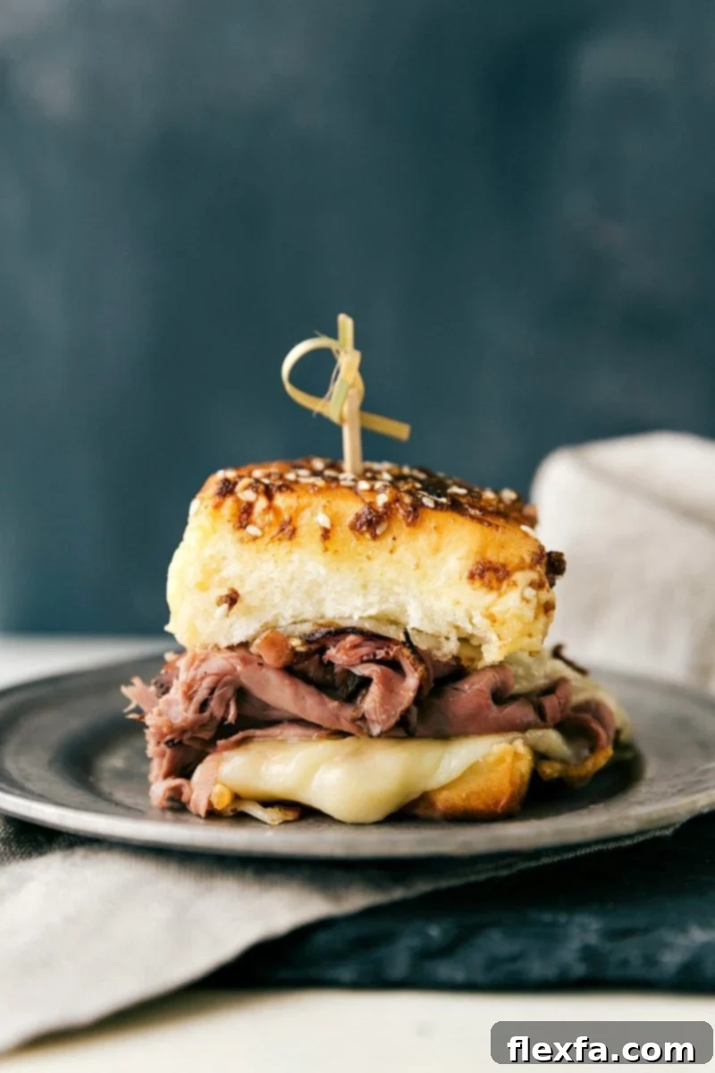 A single French Dip slider presented on a white plate, glistening with cheese and ready to be dipped into a side of au jus.