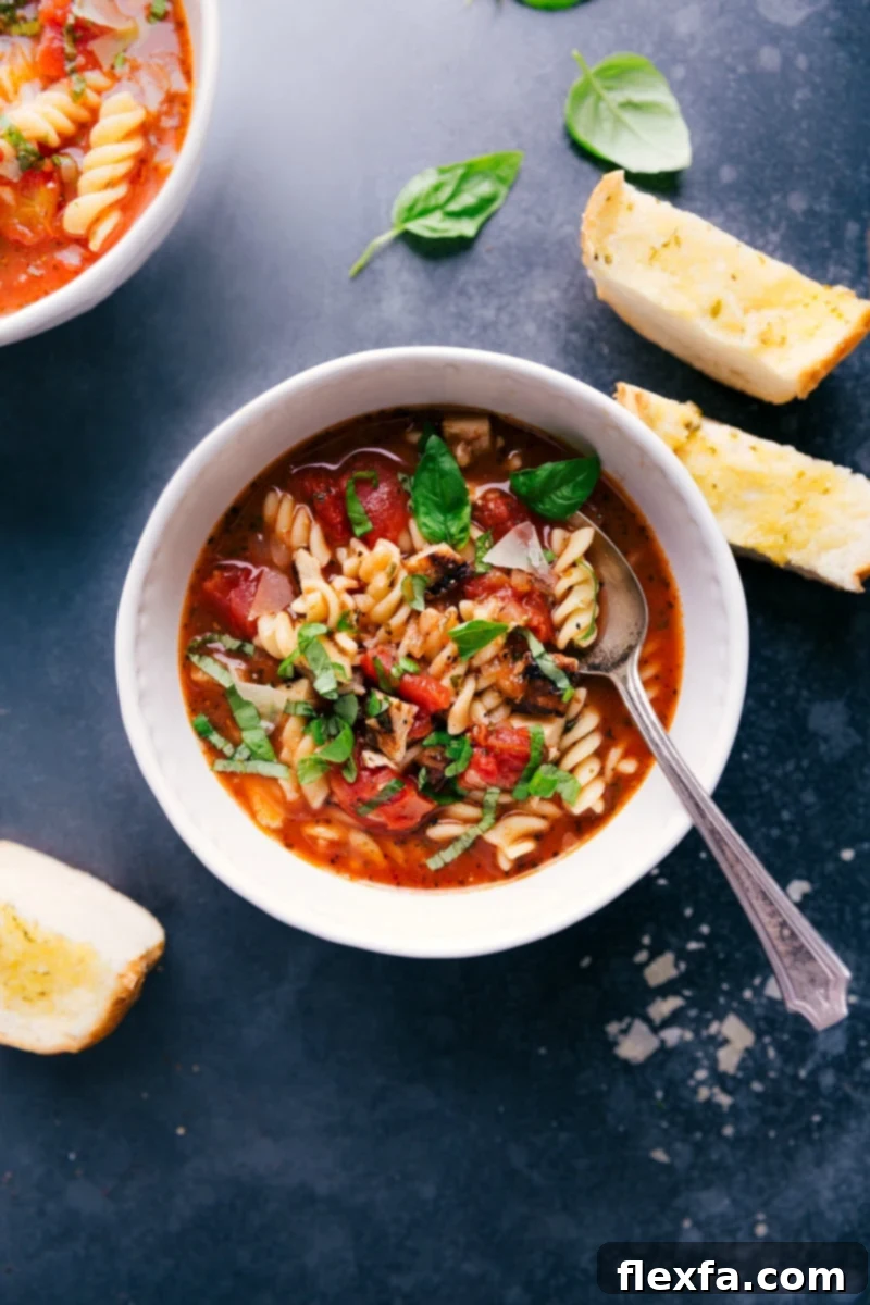 Overhead view of a beautiful bowl of Chicken Parmesan Soup, generously garnished with fresh basil leaves and shredded Parmesan cheese. It's surrounded by golden buttered garlic bread, inviting you to dive into this comforting meal.
