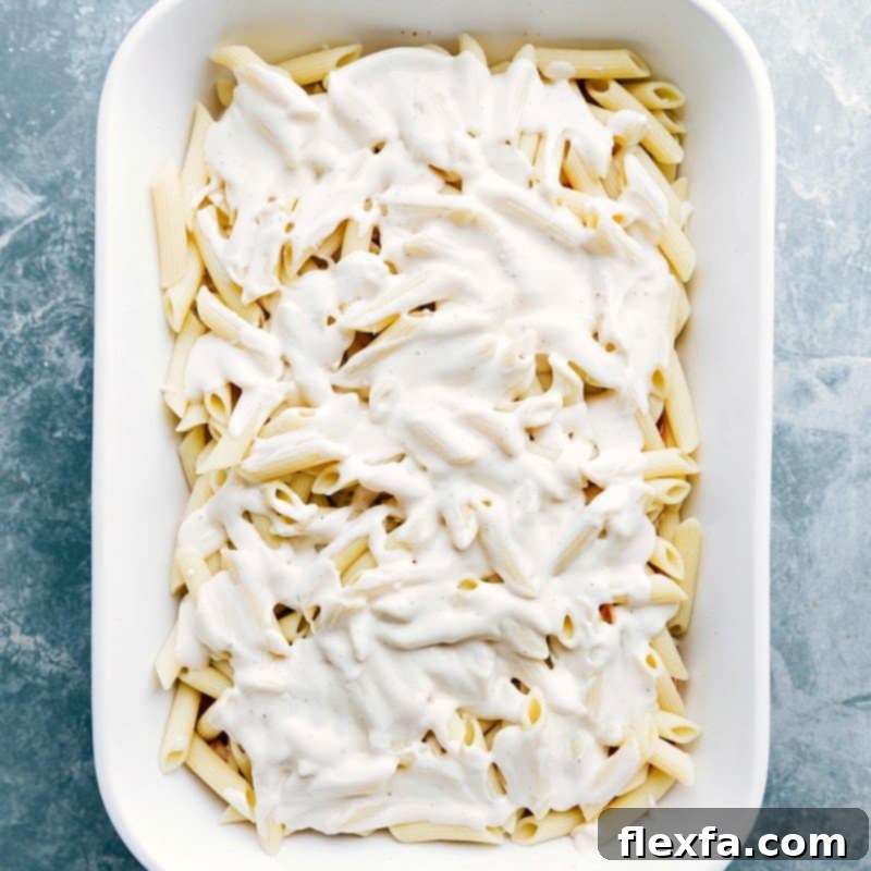 White sauce being poured over a casserole full of noodles, ready for baking.