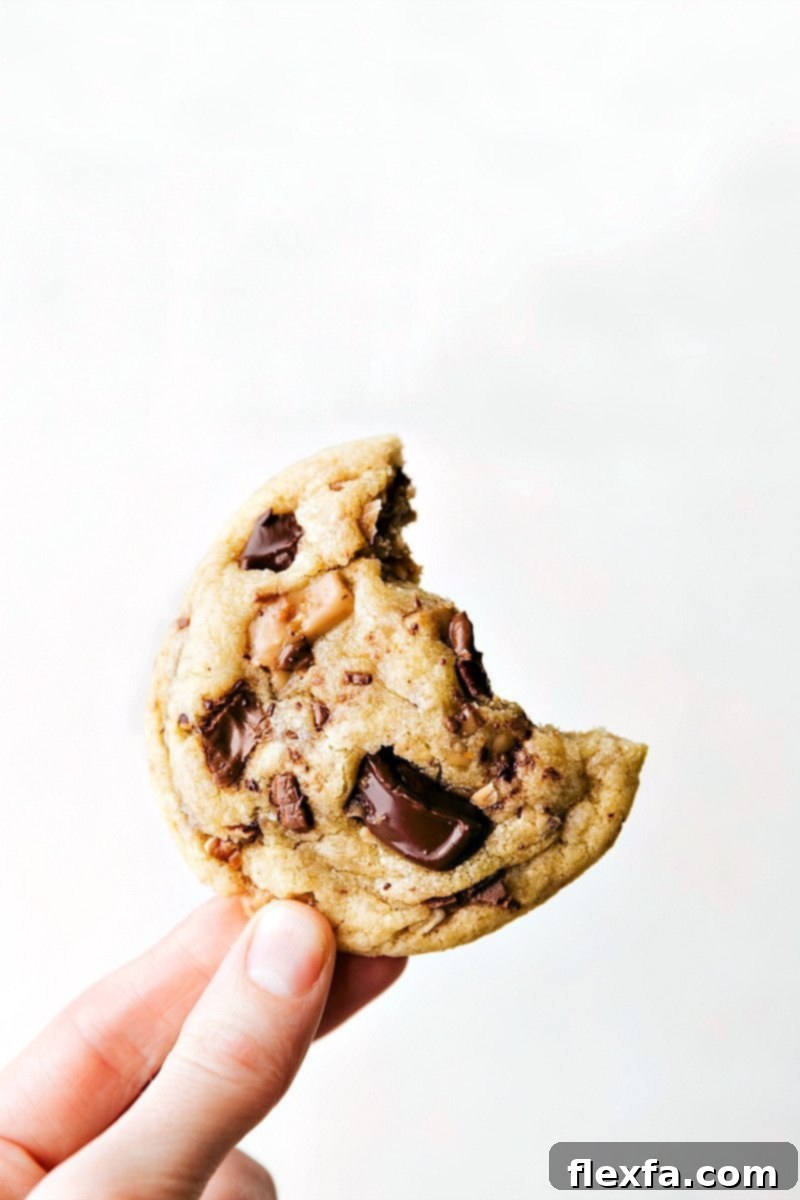 A freshly baked Toffee Cookie, golden brown with visible toffee and chocolate, held with a bite taken out, showcasing its delightful soft and chewy texture.