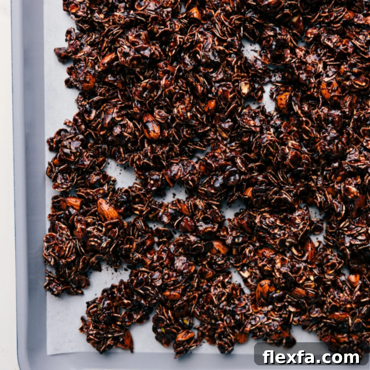 The chocolate granola mixture is evenly spread across a large baking sheet lined with parchment paper, perfectly prepared and ready to be baked to a golden, crispy perfection.