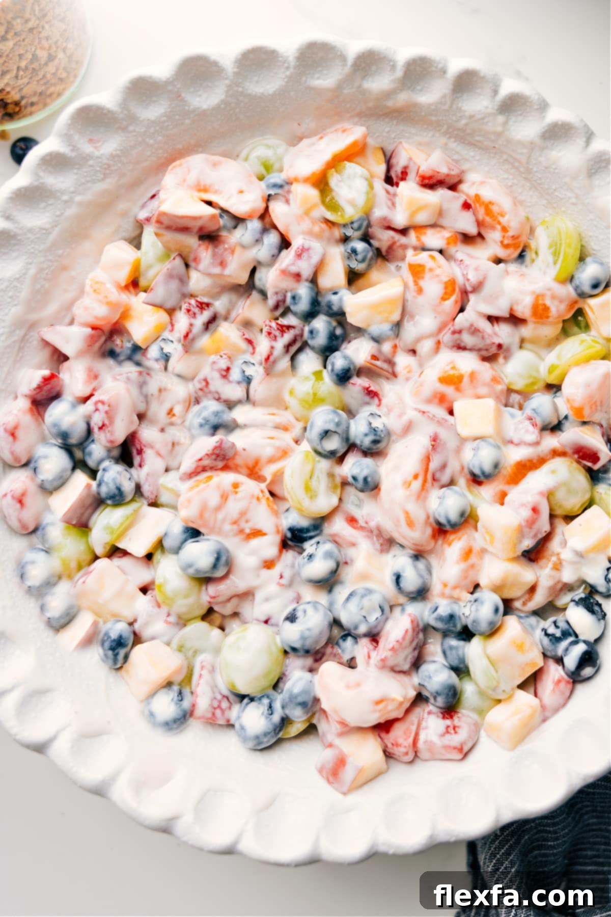 A vibrant fruit salad featuring strawberries, blueberries, grapes, mandarins, and apples, generously coated in a rich, creamy honey vanilla yogurt dressing, presented in a clear glass bowl, ready to be enjoyed.
