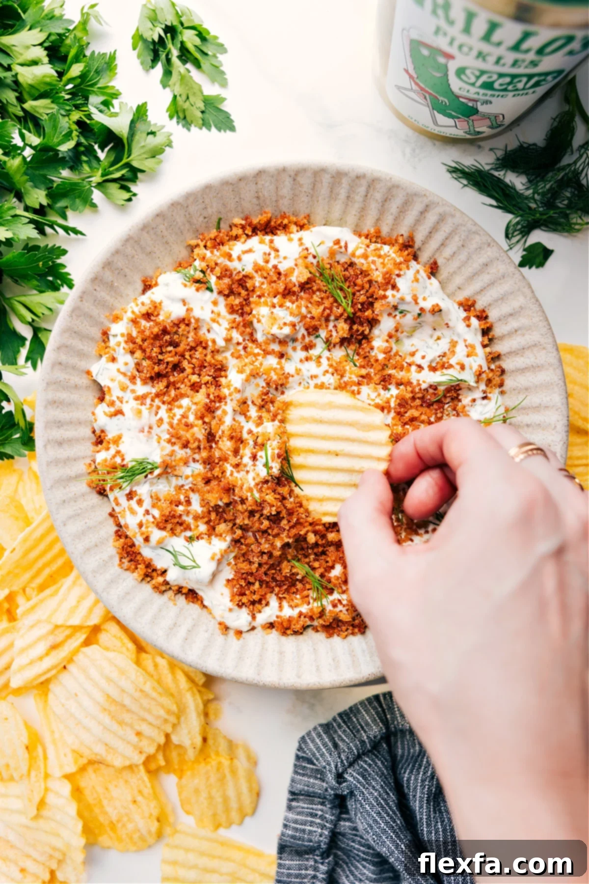 A vibrant bowl of creamy Fried Pickle Dip, generously garnished with fresh dill and a sprinkle of golden crispy panko, with a wavy potato chip poised to scoop a delicious bite.