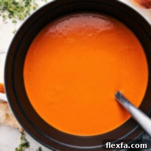 A vibrant bowl of Roasted Red Pepper Gouda Soup, garnished with a swirl of cream, fresh herbs, and a sprinkle of smoked paprika, alongside a crusty piece of bread.