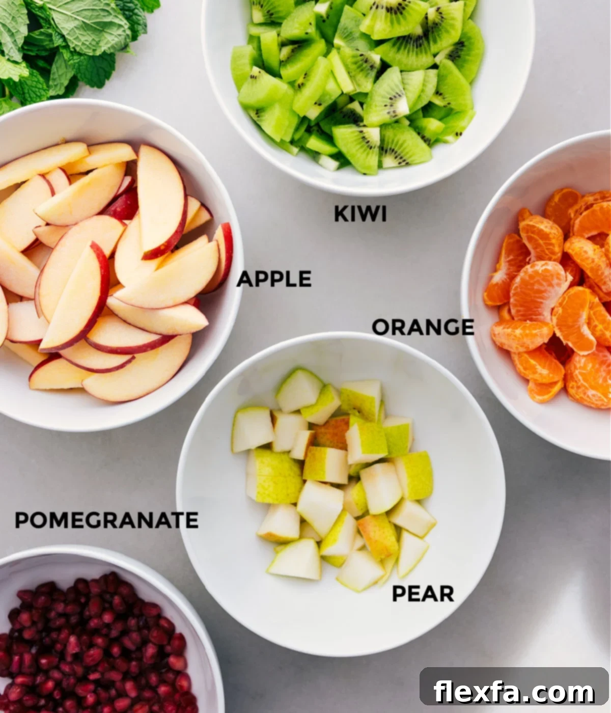 All the vibrant, fresh ingredients for this winter fruit salad are neatly prepped and arranged on a wooden board, including sliced kiwis, crisp red apples, juicy clementines, ripe green pears, and ruby red pomegranate arils, ready for assembly.