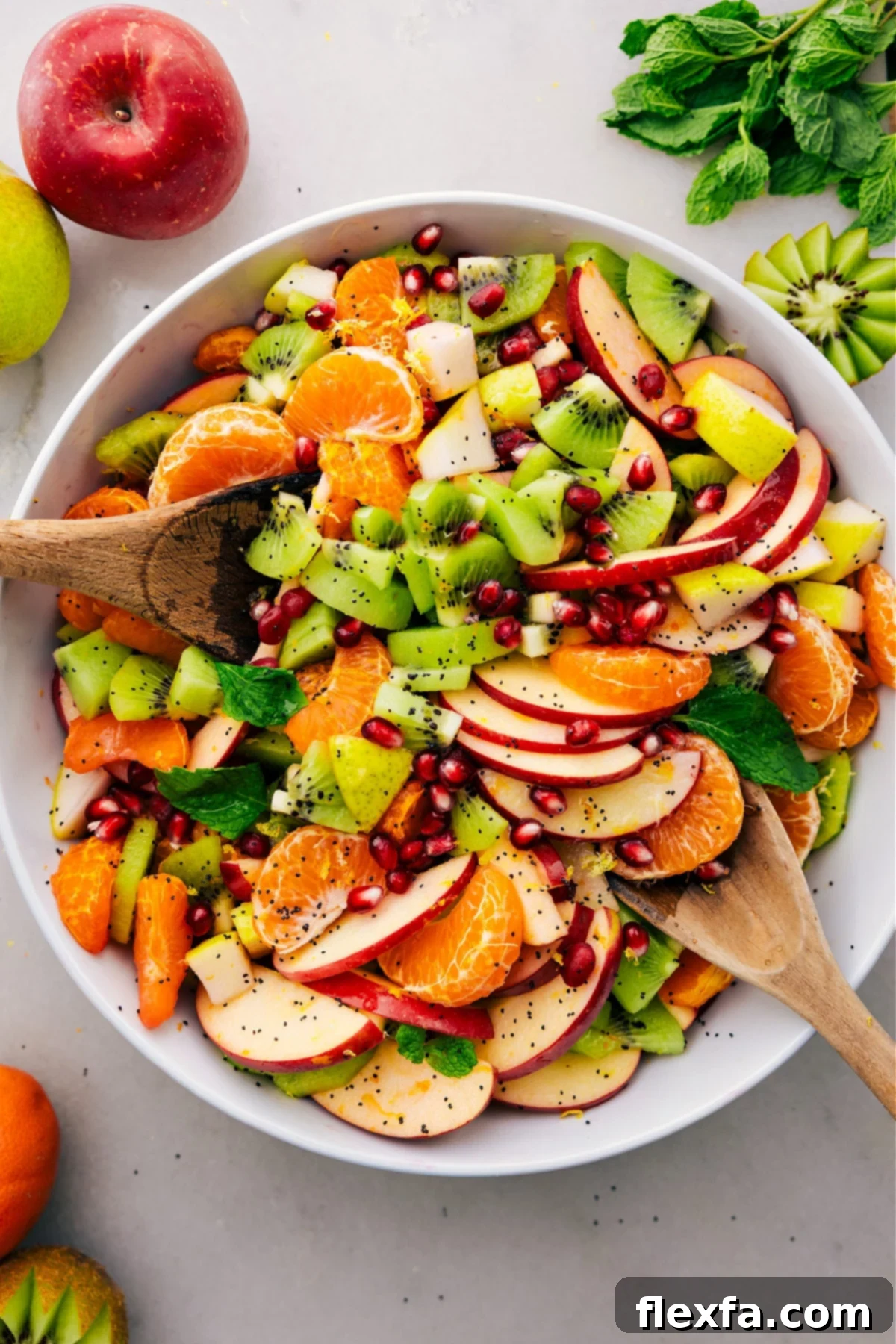 A close-up shot of a beautifully prepared Winter Fruit Salad in a rustic white bowl, highlighting the colorful array of fruits like clementines, pomegranates, apples, kiwis, and pears, perfectly glistening with the delicate poppy seed dressing.