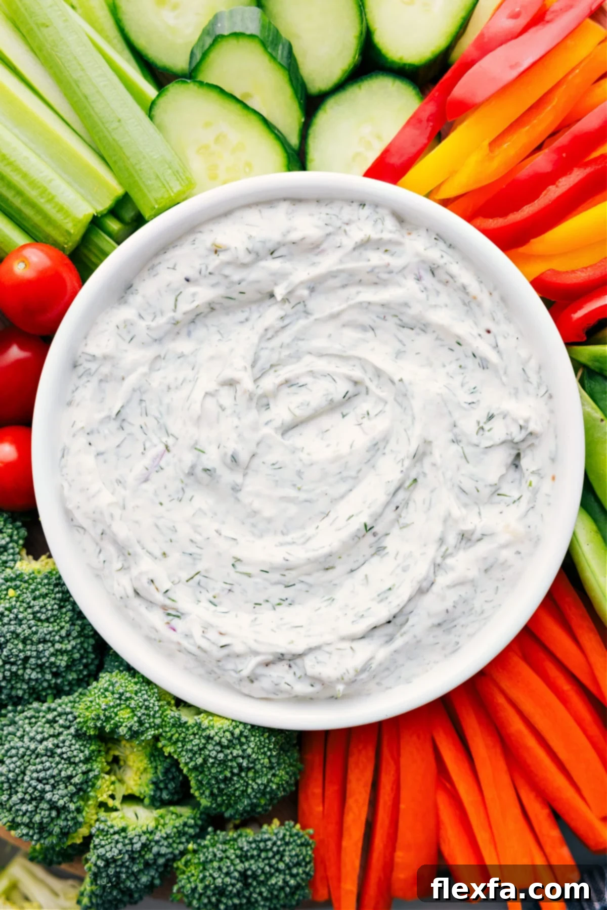 A vibrant bowl of homemade veggie dip, rich and creamy, centered on a rustic wooden board. It's surrounded by an abundant array of crisp, colorful fresh vegetables like bright red bell pepper strips, crunchy green cucumber slices, sweet cherry tomatoes, and slender carrot sticks, all perfectly arranged and ready for dipping. The scene invites a healthy and delicious snacking experience.