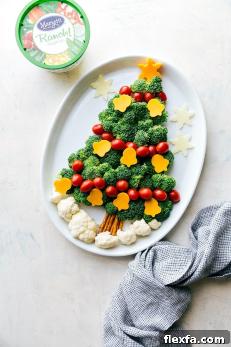 A charming and festive arrangement of green broccoli florets and bright red cherry tomatoes forming the shape of a Christmas tree. A small bowl of creamy veggie dip is placed at the base, making it a perfect and healthy holiday snack or appetizer.