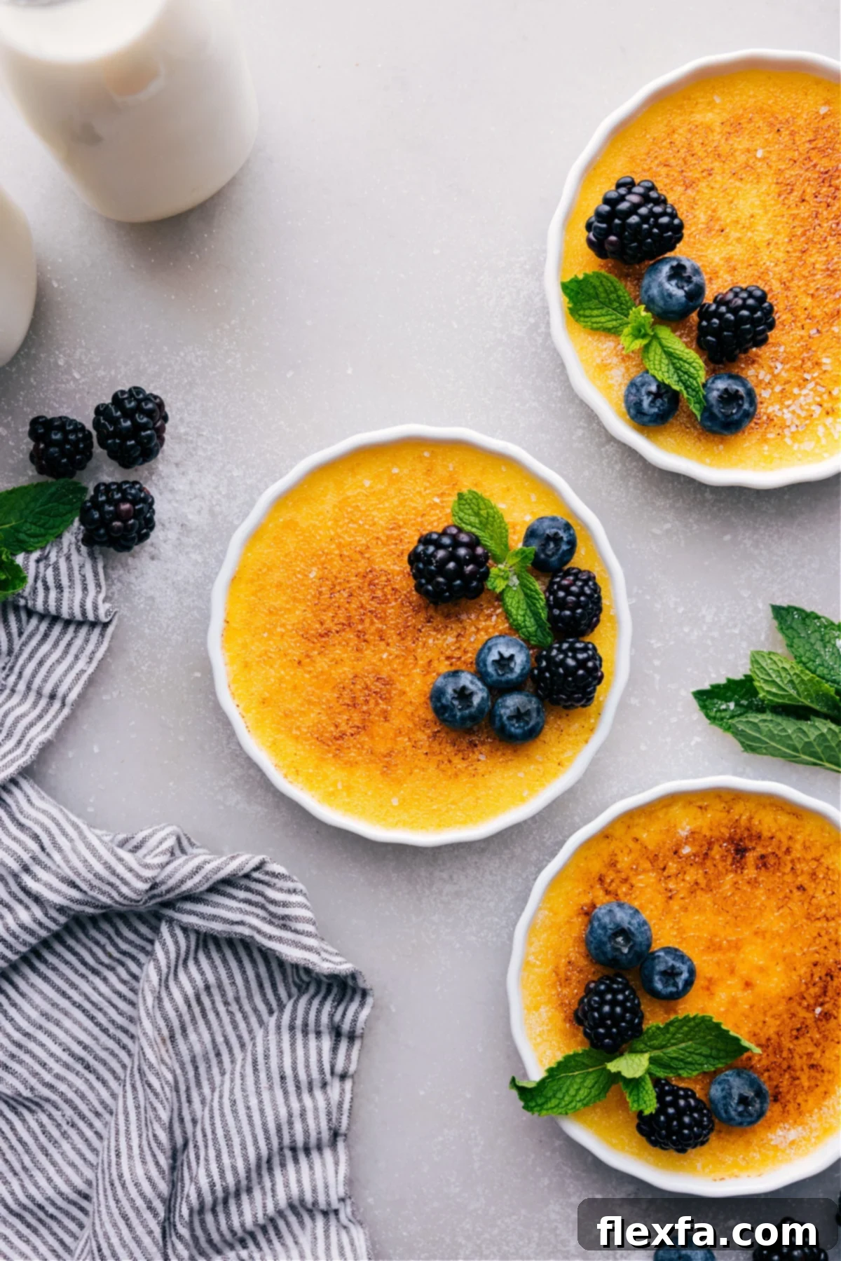 Golden crème brûlées with a crisp sugary top, garnished with vibrant fresh berries and delicate mint leaves, presented beautifully on a white serving platter.