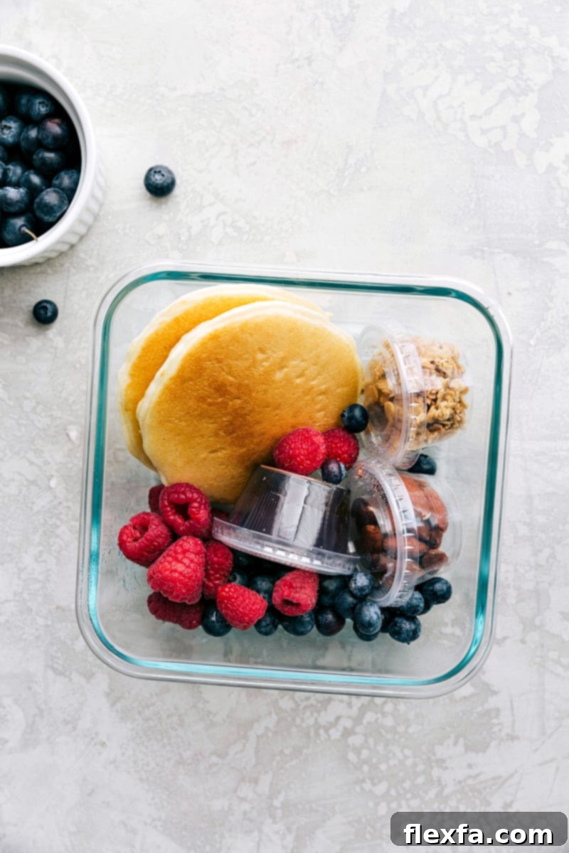 Pancakes, syrup, granola, nuts, and fruit packed in containers, ready to go for a wholesome kids school lunch.